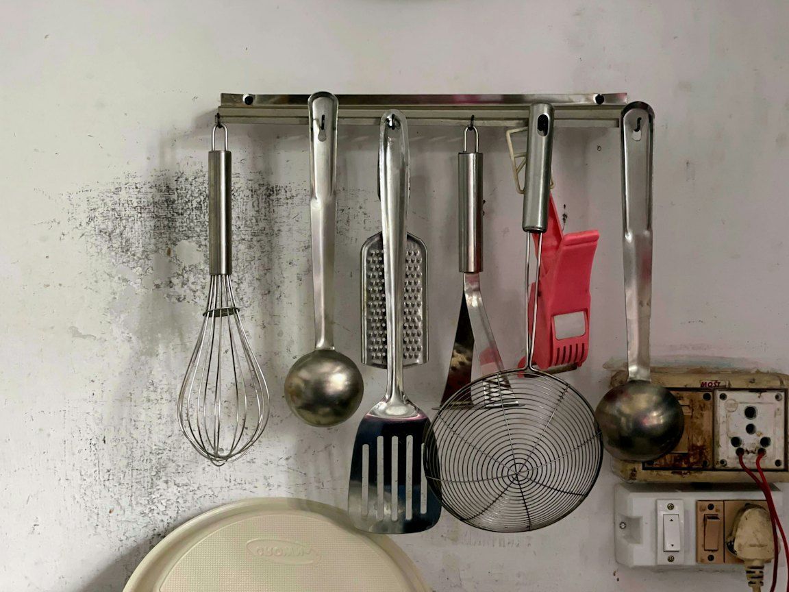 A whisk, two ladles, a grater, a turner and a skimmer hang from a metal hook rail fixed to a pale wall.