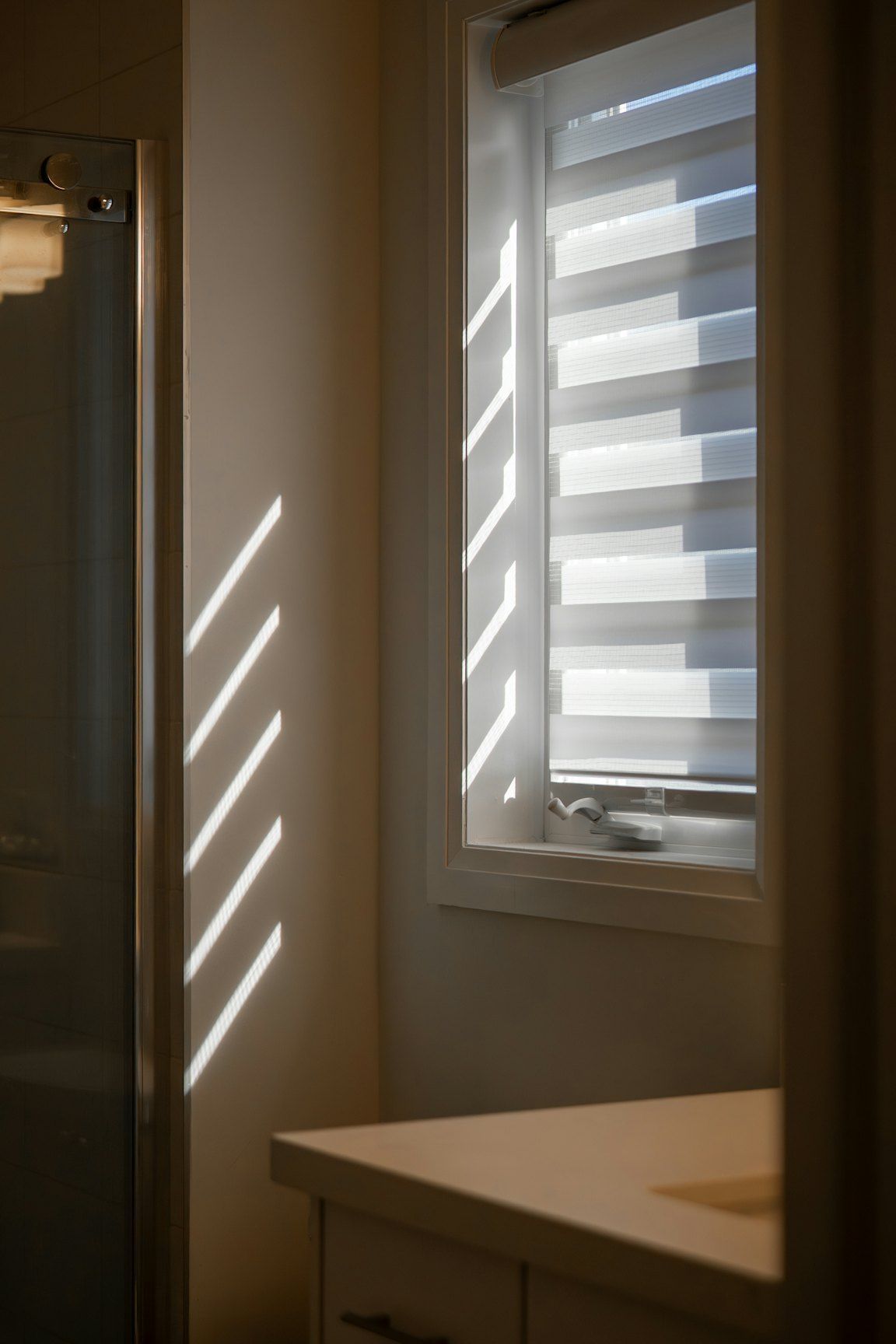 A blind with alternating sheer and opaque bands on a small window next to a shower screen, with stripes of sunlight on the wall.